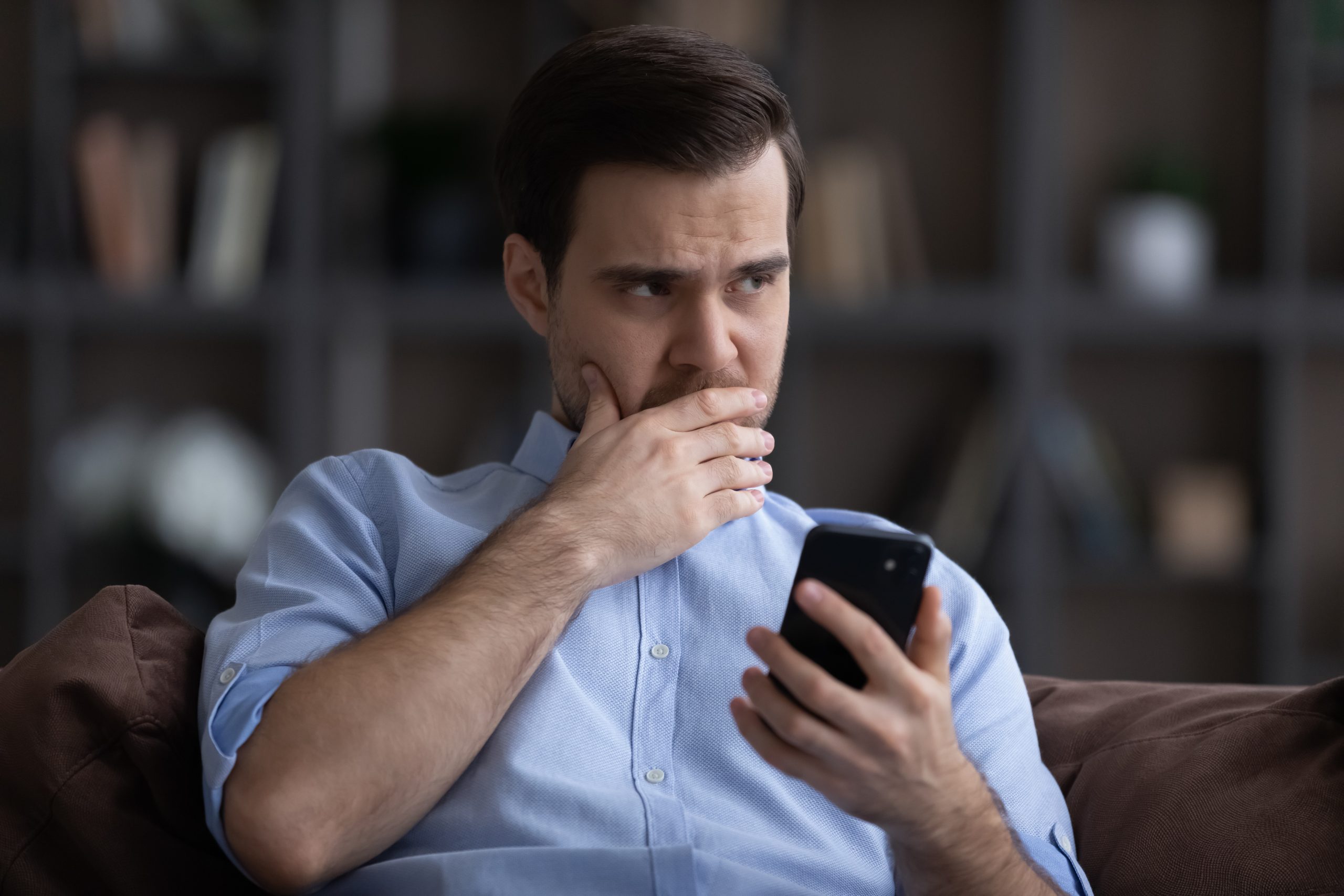 Doubtful,Young,Man,Sit,On,Sofa,Hold,Phone,Look,Aside